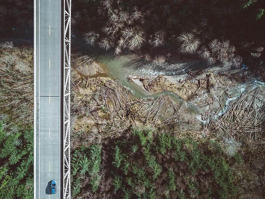 An aerial view of a bridge over a winding river, surrounded by dense forests. A single car is seen driving on the road above, with a contrast of dark tree cover and lighter areas near the water.