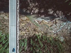 An aerial view of a bridge over a winding river, surrounded by dense forests. A single car is seen driving on the road above, with a contrast of dark tree cover and lighter areas near the water.
