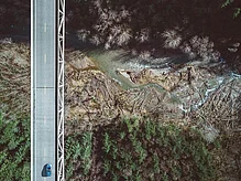 An aerial view of a bridge over a winding river, surrounded by dense forests. A single car is seen driving on the road above, with a contrast of dark tree cover and lighter areas near the water.